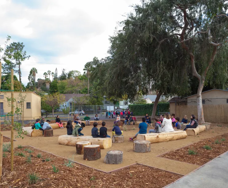 Elementary school students sitting on logs in a schoolyard surrounded by plants and trees