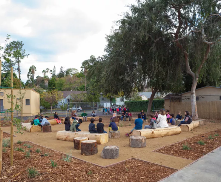 Elementary school students sitting on logs in a schoolyard surrounded by plants and trees