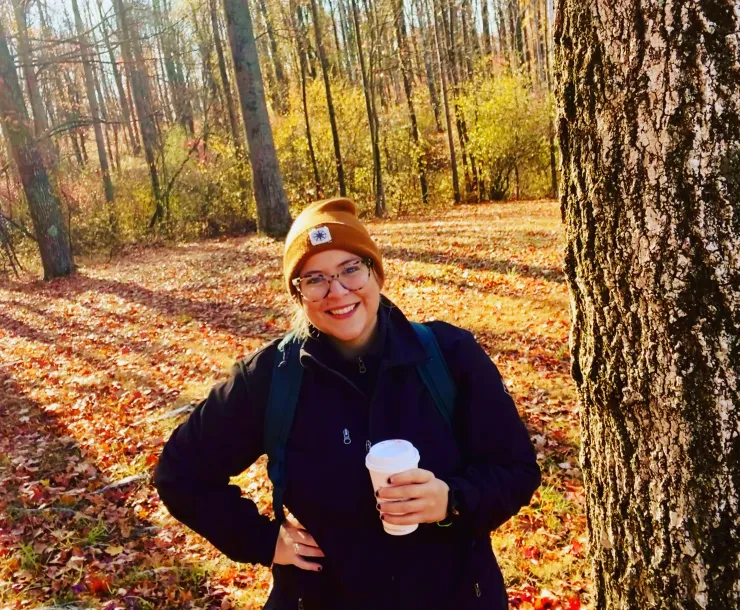 A woman with glasses and a brown winter hat stands outside in the woods. There are many autumnal leaves on the ground. She is smiling and holding a beverage in one hand, and the other hand is on her hip. She is wearing a black coat.