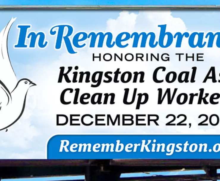 Billboard against a blue sky which says "In Remembrance, Honoring the Kingston Coal Ash Clean Up Workers"