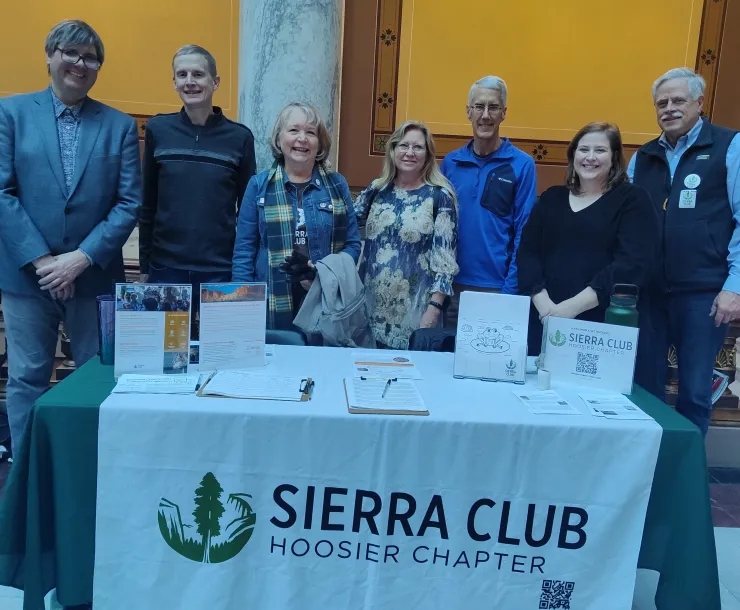 A groups of seven people standing behind a table indoors at the Statehouse. On the table is a white tablecloth with the green and black Sierra Club Hoosier Chapter logo. Everyone is smiling for the photo.