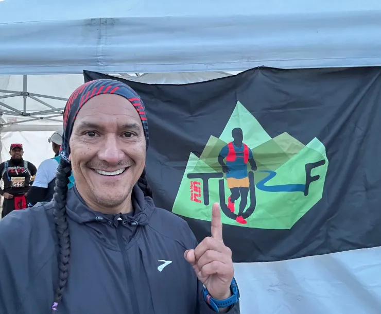 A man smiling for the camera outdoors at a running event. There is an event tent behind him and a couple of other people are in the background. He has a black wind jacket and long dark brown/black hair in a plait. He is pointing at a black banner with a green TURF logo which stands for Trail and Ultra Running Fanatics.
