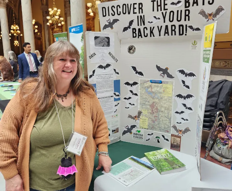 A person with a green top and an orange cardigan, long brown/gray hair with bangs and smiling, tabling at an indoor event at the Statehouse. On the table is a display: Discover the bats in your backyard.