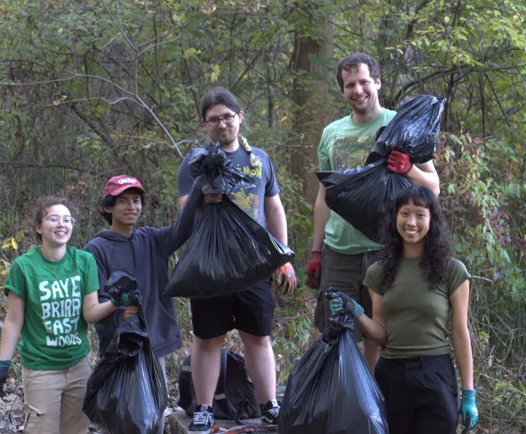 A group of five people outdoors with trees behind them. They're holding black trash bags. Some are wearing gloves, and one is wearing a green t-shirt with Save Briar East Woods in white letters.