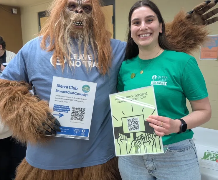 A person in a green t-shirt with the words Sierra Club organizer stood next to a person dressed as a very hairy mascot with a blue t-shirt. The mascot is holding a sign for a Sierra Club Beyond Coal campaign with a QR code, and the person in the green t-shirt is holding a sign saying to oppose Indiana senate bill 1007
