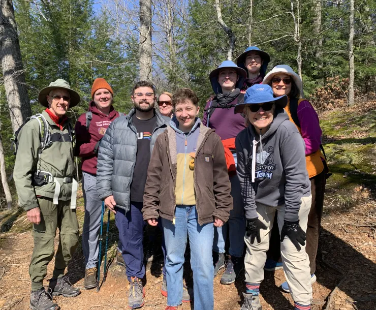 A group of nine people stood outdoors on a trail in a woods. It is a sunny day and there are pine trees with green needles behind them. Everyone in the group is smiling.