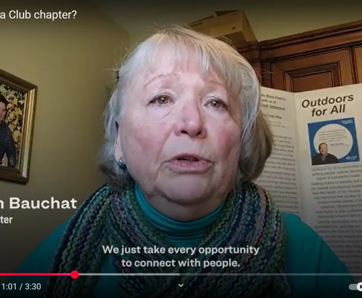 A screenshot from a video of a person sitting at home with a board behind them saying "outdoors for all" and a family photo also on the wall. The text on the video says Marilyn Bauchat and the subtitles say "We just take every opportunity to connect with people"