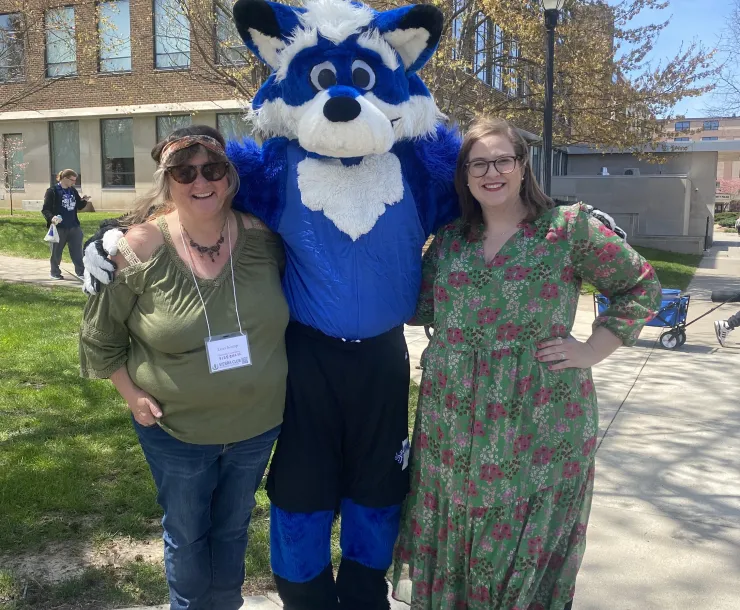 Two people stood either side of a blue and white furry animal mascot. They are both smiling. The person on the left is wearing a green top and blue jeans, and the person on the right is wearing a green dress with purple and white flowers. There is a building behind them and trees and blue sky.