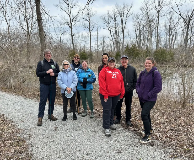 A group of 8 people stood outdoors on a gravel trail. There is a large pond behind them as well as bare trees, and brown leaves on the ground. They are dressed for a cool day.