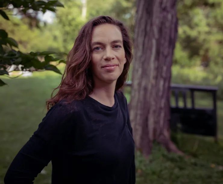 A woman with long brown hair stood outside in her yard - the background of trees and grass is slightly blurred in contrast with the person in the foreground. She is standing side on but looking towards the camera and is wearing a dark top with long sleeves.