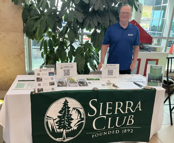 A person tabling indoors. They are wearing a blue shirt and are stood behind a table with a green and white Sierra Club tablecloth. There are leaflets and other sources of information on the table.
