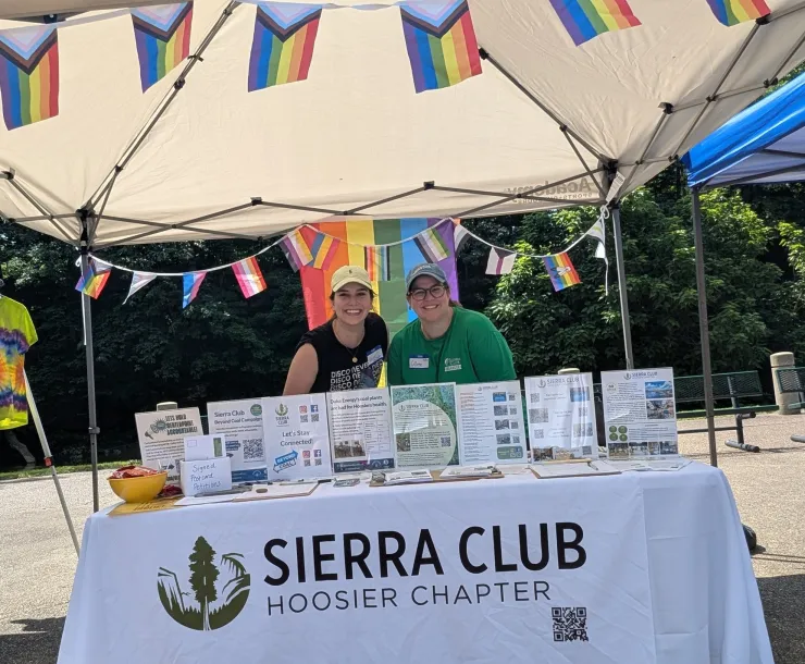 Two peope stood behind a table outdoors. The table has a Sierra Club Hoosier Chapter banner and Pride flags are hanging from the canopy aboe the table. The table has materials and displays on it.