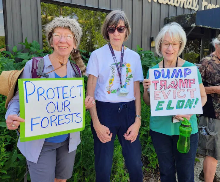 Three people standing outside a Hoosier National Forest building. Two of them are holding signs - one says Protect our Forests, the other Dump Trump Evict Elon!
