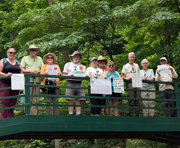 A group of ten people on a bridge with green trees in the background. Some are holding signs which say things like 'take a stand for public land' and 'save our forests'