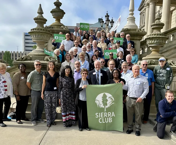 A diverse group of people are gathered on the steps of the state capitol in Lansing Michigan holding a Sierra Club Michigan Banner