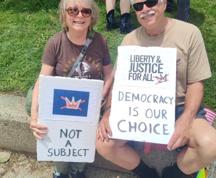 Two people sat at a rally - they are holding signs. One has a crossed-out crown with the words "not a subject" and the other says Librety & justice for all - Democracy is our choice.