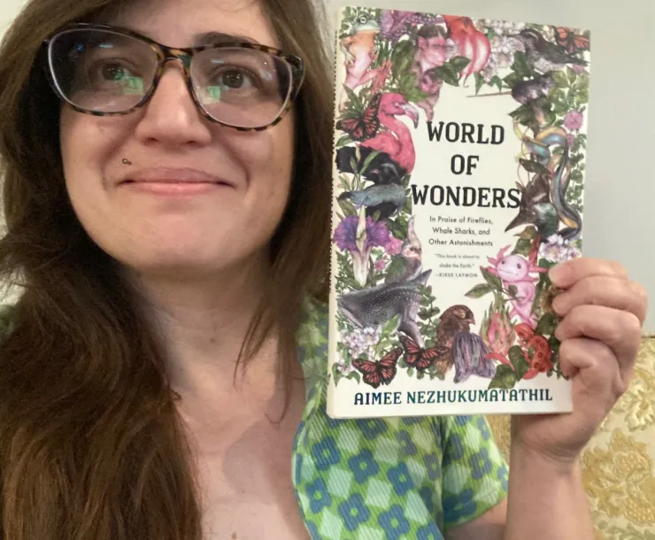 A person with long brown hair and glasses indoors, smiling and holding a book called World of Wonders which has a colorful illustration of many animals and flowers on the cover.