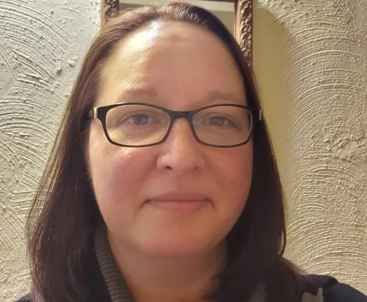 A head and shoulders shot of a woman with brown hair and dark glasses, indoors with an off-white wall behind her.