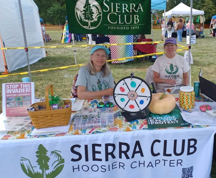 Two people tabling outdoors. There is a green Sierra Club banner hanging behind them, and a white table cloth which says Sierra Club Hoosier Chapter and has a green tree logo on it, on the table. Both of the women at the table are wearing shirts with the Sierra Club logo, and baseball caps. On the table there is information and handouts.