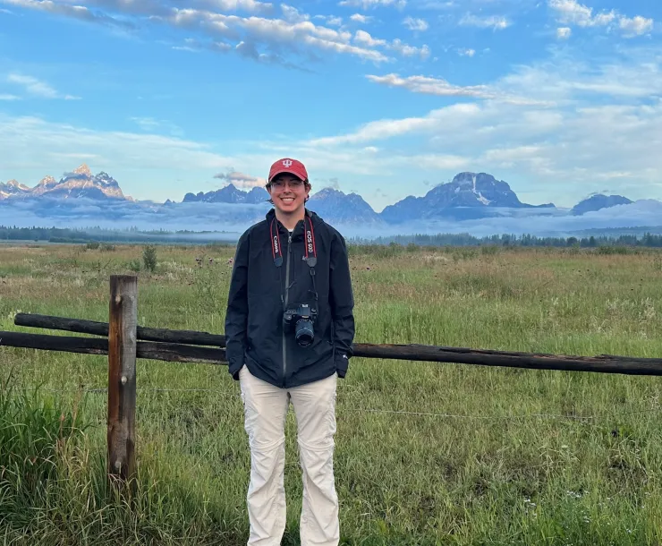 A person standing next to a large expanse of grasslands with mountains shrouded in mist behind them. The sky is bright blue, scattered with light white and gray clouds. The person is wearing a red IU cap, a black jacket, white pants, and has a camera around their neck. They are smiling.