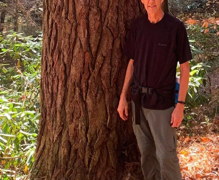A man with short gray hair standing next to the base of a wide tree. He is smiling, and wearing a black tee and gray pants. 