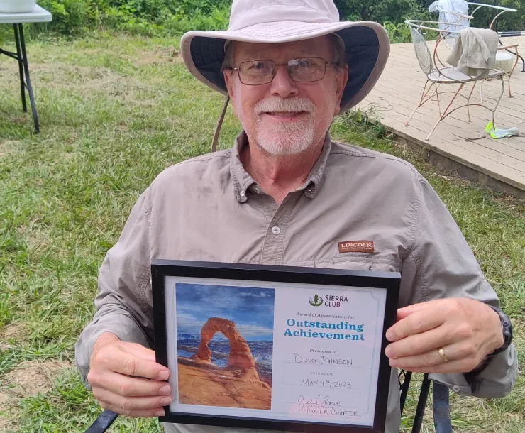 A man with a neat gray beard, glasses, and a tan hat and shirt, sitting in a chair in a yard. He is smiling, and holding a framed award which says Award of Appreciation for Outsanding Achievement presented to Doug Johnson by Julie Lowe, Sierra Club Hoosier Chapter on May 9, 2023.