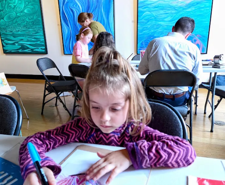 A young child with brown hair and a stripy purple long sleeved tee, drawing on paper with colored pens. She is sat at a table indoors. There are some other people sat at a table behind her doing similar activities, and paintings on the wall.