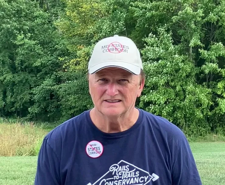 A man outdoors with green grass and green trees in the background. He has a white cap stating No Mid-States Corridor, a blue t-shirt, and a button also against the Mid-States Corridor