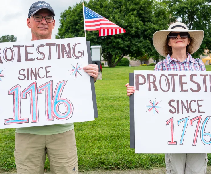 two people hold up pro-democracy signs with an American flag in the background