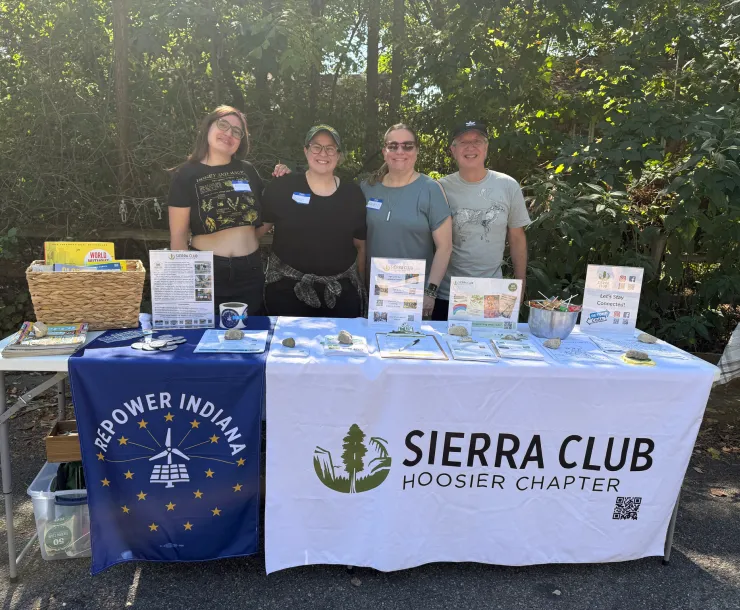 Four people tabling outdoors on a street. There are trees behind them. They are smiling for the photo. On the table there is a white tablecloth with the Sierra Club Hoosier Chapter logo of black text and a green tree graphic, and a smaller blue tablecloth which says Repower Indiana with a white and yellow logo of a wind turbine and solar panels surrounded by a circle of stars. On the table there are books, petitions, stickers, and other info about the Sierra Club.