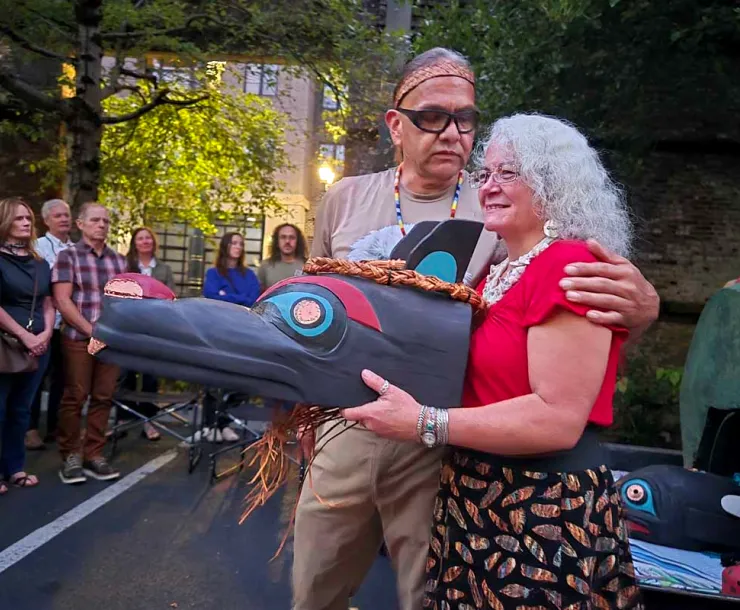 A man has his arm around a woman who holds a large cedar mask in front of her body