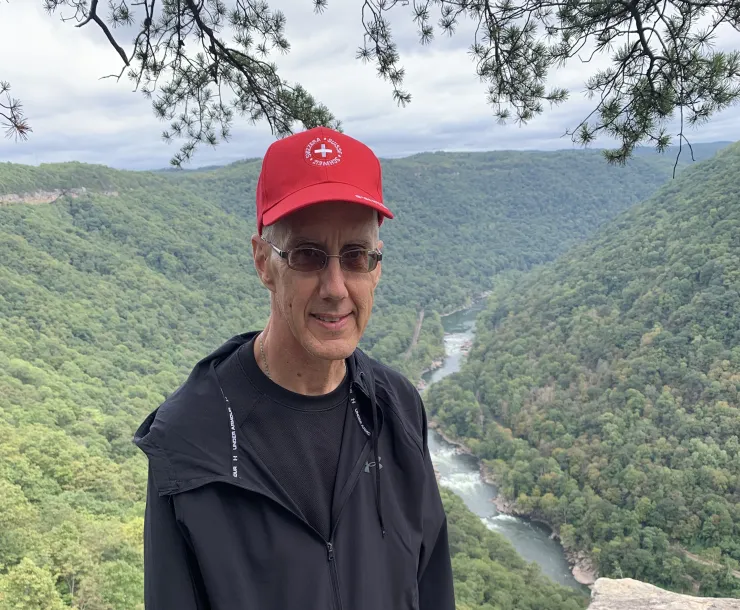 A man with a red cap and black jacket, and sunglasses standing on a rocky outpost with a forested valley behind him.