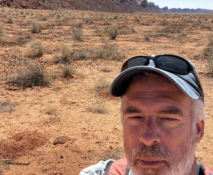 A head and shoulders selfie of a person with a gray cap and top, wearing a backpack with orange and black straps, outside in a desert-looking park. The sky behind him is a hazy blue and there are hills or small mountains behind him, and small dark green plants growing from the ground.
