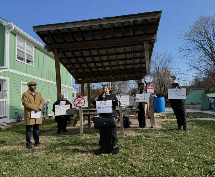Five people stood outdoors in a small neighborhood park. One person is standing forward of the others and is speaking through a microphone. There is a sign on the microphone stand saying NO data center. The other people in the photo are all holding signs too. There area  few trees in the background which have shed their leaves. The sky is blue.