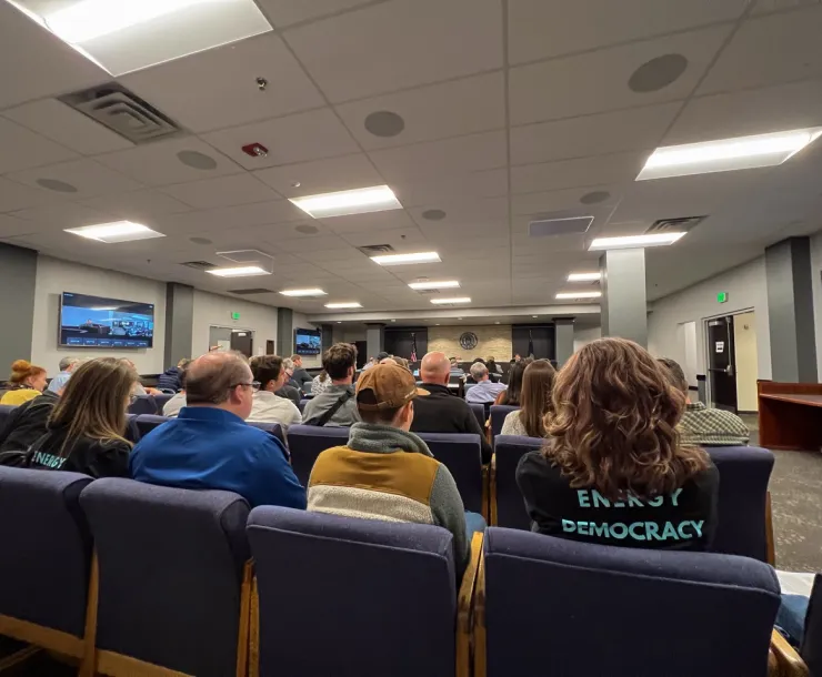 Picture of people sitting in chairs in a public hearing room at the Idaho PUC, one person with a shirt that says Energy Democracy on the back