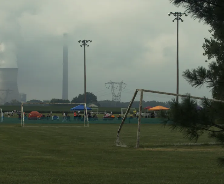 Coal plant towers billowing out smoke in background. In the foreground is a sports field with soccer goals with some players gathered at the far end. The sky is a smokey gray.