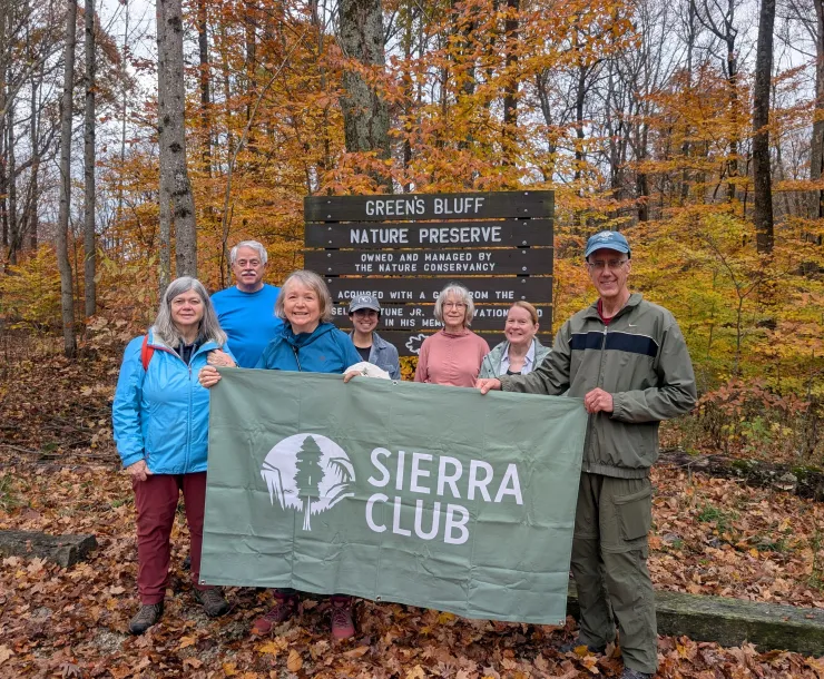 A group of 7 people at a trail head. They are holding a green Sierra Club banner and dressed for the cooler weather. The leaves on the ground and on the trees are brown and yellow.