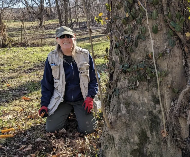 A person kneeling down by the base of a large tree. They're wearing gloves and have a tool for removing invasive species in their hand. They're wearing a green cap and dark sunglasses. The grass is green and there are other trees in the background. They are smiling.