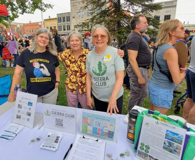 Three people tabling at a No Kings rally. They are in a park and there are buildings behind them as well as some trees. There are crowds of people in the background, and some folks are holding signs. The three people posing for the photo are standing behind a table which has Sierra Club literature and signage.