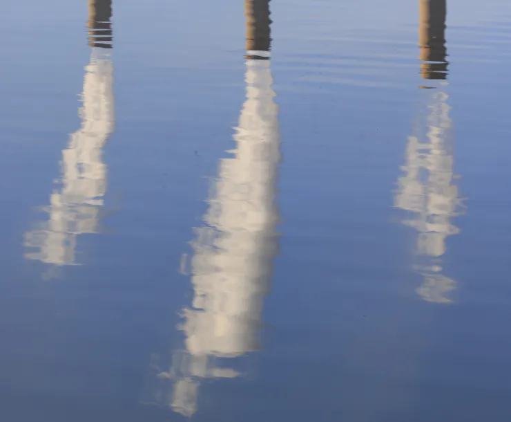 Three smoke stacks reflected in water. The smoke is white and the stacks are brown/gray. The water is blue.