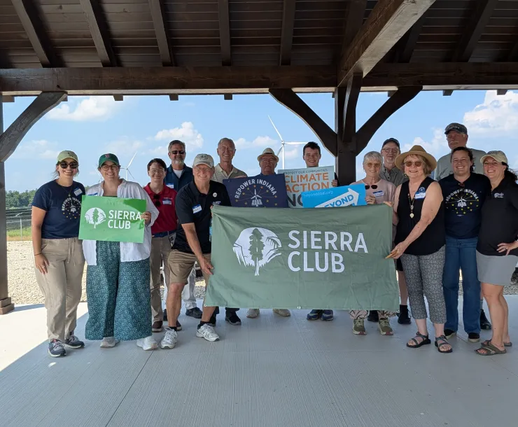 A group of fourteen people standing outside. Some of them are holding signs and banners saying "Sierra Club" and "Repower Indiana". The sky behind them is blue with small fluffy clouds, and a couple of wind turbines are also in the background.