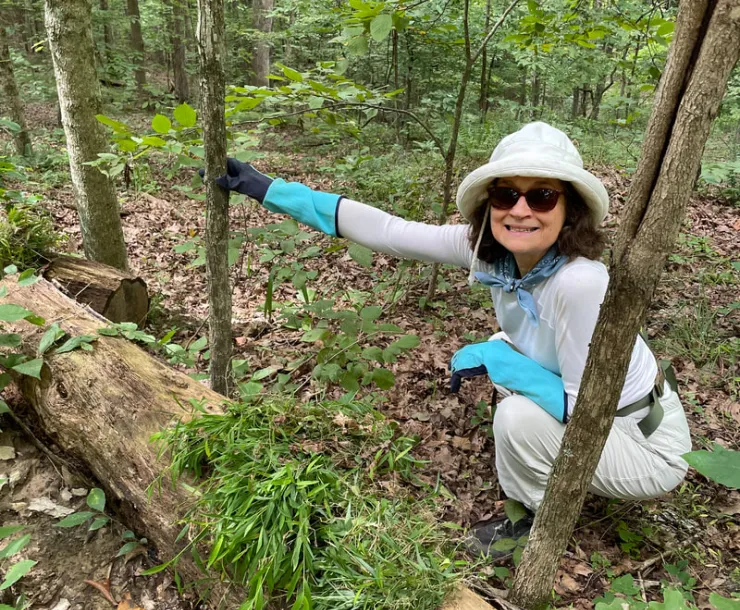A person kneeling down in the woods holding onto a sapling with one hand. They're wearing gloves, a white cap and dark sunglasses. They are smiling.