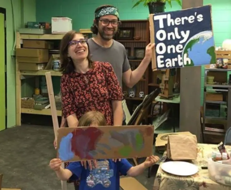 Two adults and one child in an art studio workshop. The tallest adult is holding a painted sign saying There's Only One Earth with a painting of the Earth on it. The child is holding up painted cardboard in front of their face. The two adults are smiling.