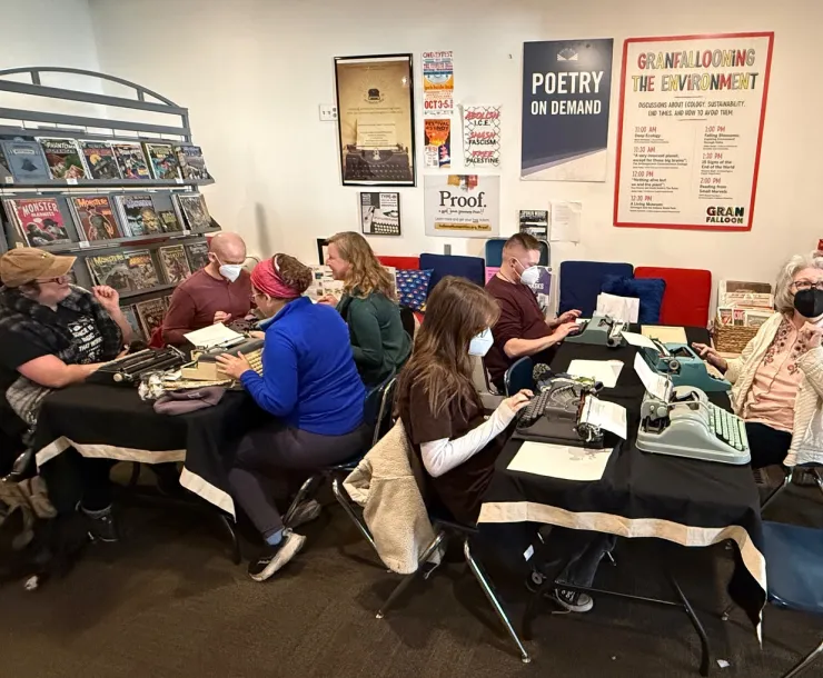 A group of seven people sitting at tables with typewriters. Several of the people are wearing masks to protect them from airborne viruses. Everyone looks happy - some are in conversation, some busy typing. The room they're in has white walls with posters and some shelves with comics and graphic novels. One of the people has a dog sitting by them.