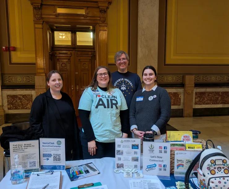 Four people tabling indoors. There is lots of information on the table including flyers and petitions. One of the people is wearing a shirt saying I heart clean air. Another is wearing a shirt saying Repower Indiana. All four people are smiling.