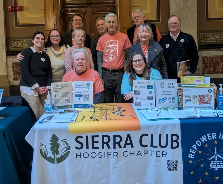 A group of 11 people indoors sitting or standing behind a table. On the table are petitions to sign, a light orange poster with a map of Indiana and markers, and other information about the Sierra Club. There is a white banner with a Sierra Club Hoosier Chapter green tree logo and black writing on the table. The people in the photo are smiling.