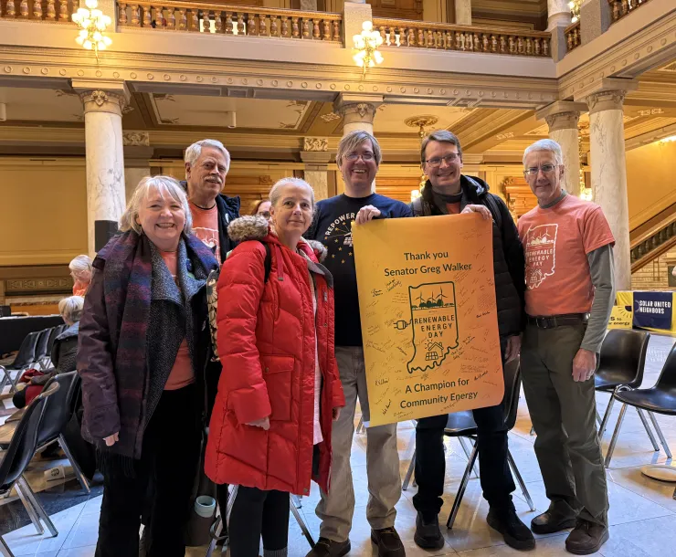Six people standing indoors at the Statehouse. There are pillars behind them and chairs spread out in the room for attendees. Two of the people are holding up a yellow poster with an outline of the Indiana map on it. The poster says Thank you Senator Greg Walker. A champion for Community Energy. The people in the photo are smiling.