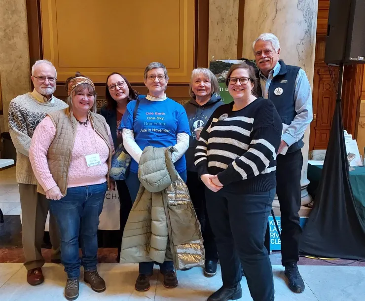 Seven people of varying ages standing indoors at the Statehouse. They are all smiling for the photo.