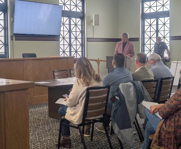 A person in a salmon pink jacket standing behind a podium by the side of a room giving a presentation on a screen. There are people sitting on chairs watching and listening.