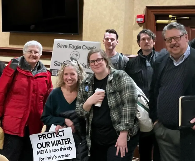 A group of 7 people indoors at an official building. Some are holding signs saying PROTECT OUR WATER - META is too thirsty for Indy's water, Save Eagle Creek, and MAKE META RECYCLE THEIR WATER.